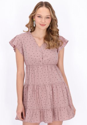 Nuori nainen aaltoilevilla hiuksilla, pinkki pilkullinen mekko, jossa liehuvat hihat ja kerroksittainen helma, hymyilee kameralle.