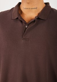 Bruine polo met een textuurstof, twee knopen aan de voorkant en een korte kraag. Bevat een kleine opening aan de voorkant en subtiele stiksels.