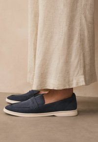 Navy blue suede slip-on shoes with a flat white sole and decorative stitching. A soft, textured finish complements casual attire.