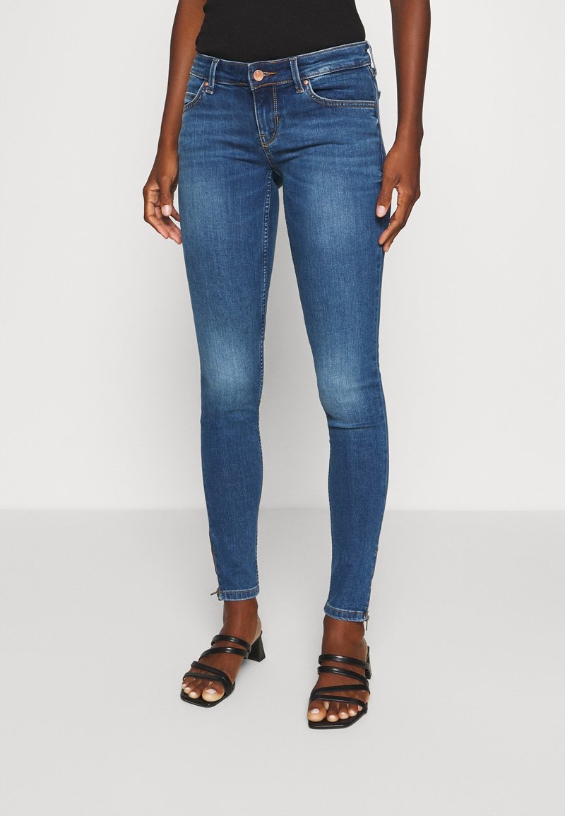 Jeans de mezclilla azul ajustados con cremalleras en los tobillos, llevados por una persona de pie, combinados con sandalias de tacón negras con tiras sobre un fondo liso.