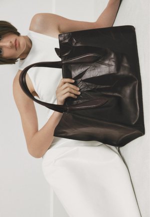Bruine leren tote bag met twee handvatten, met een gladde textuur en een lichte kreukeling. Tentoongesteld tegen een neutrale achtergrond.