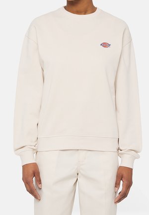 Persoon draagt een crèmekleurige trui met lange mouwen en een ronde hals, met een klein meerkleurig logo op de linkerborst.