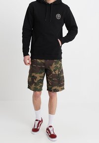 Felpa nera con tasca a marsupio e lacci, abbinata a pantaloni cargo mimetici. Calzature: scarpe da ginnastica rosse e bianche. Calze bianche visibili.