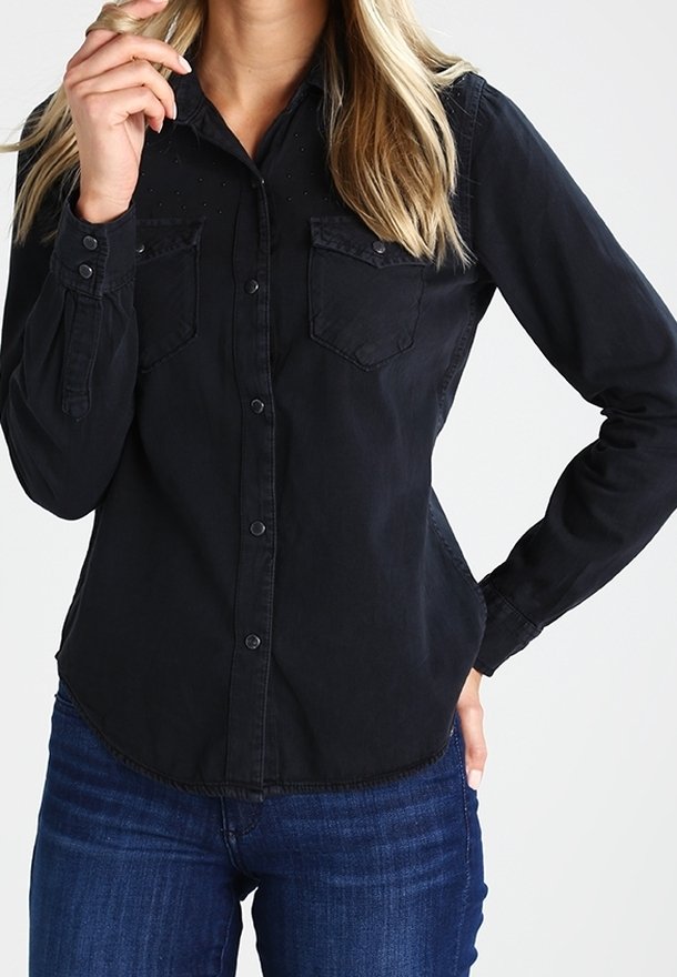 Femme portant une chemise noire à boutons avec deux poches poitrine et un jean bleu, une main près du visage et l'autre sur la hanche.