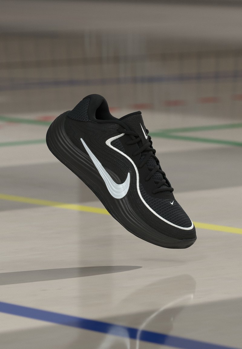 Μαύρο αθλητικό παπούτσι με λευκό λογότυπο Nike Swoosh, στον αέρα πάνω από το δάπεδο γυμναστηρίου με σημάνσεις με χρωματιστές γραμμές για αθλήματα.