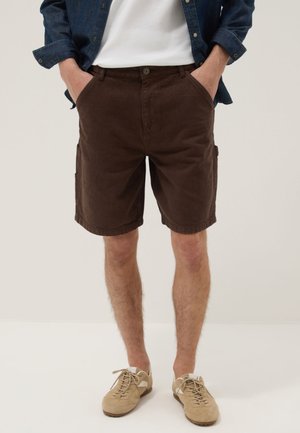 Man draagt donkerbruine korte broek met zijzakken, een wit T-shirt, een spijkerjasje en lichtbeige veterschoenen.