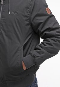 Czarna kurtka bomberka na zamek błyskawiczny, z prążkowanymi mankietami i kołnierzem. Posiada naszywkę z logo na rękawie oraz boczne kieszenie. Gładka tekstura.