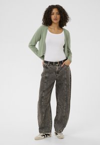 Lichtgroene cropped cardigan over een wit tanktopje, gecombineerd met zwarte high-waisted jeans met een acid-wash en casual witte sneakers met zwarte accenten.