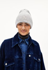 Grijze geribbelde gebreide beanie met subtiele spikkels, voorzien van een geborduurd logo. Gecombineerd met een marineblauwe corduroyjas en een donkere kraaglaag.