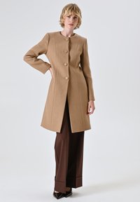 Cappotto di lana color tan con silhouette dritta, colletto rotondo e sei bottoni; abbinato a pantaloni marroni e scarpe nere a punta.