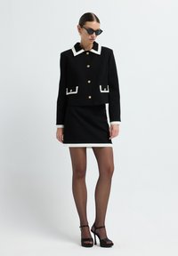 Schwarze, kurze Jacke mit weißen Akzenten und goldenen Knöpfen, kombiniert mit einem schwarzen Minirock mit weißem Saum. Das Model trägt schwarze Cateye-Sonnenbrillen.