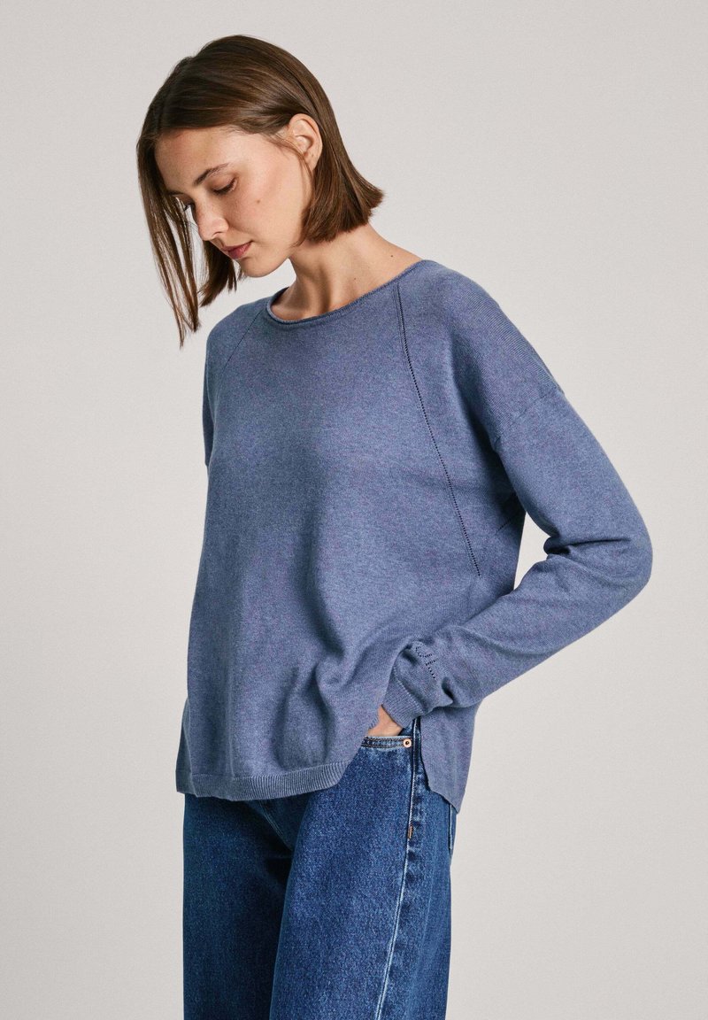 Modra pletena jopica z okroglo ovratnikom, dolgimi rokavi in stranskimi razporki, v kombinaciji z visokimi kavbojkami. Mehak material, sproščen kroj.