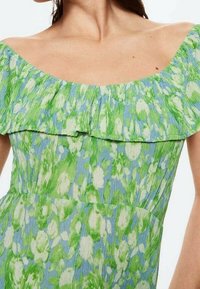 Robe florale avec un décolleté à volants, sans épaule. Fabriquée dans un tissu léger avec un motif vert et bleu. Finition texturée.