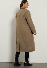Donna che indossa un lungo cappotto color cammello, pantaloni neri e stivaletti neri con tacco, in piedi contro una parete chiara, girata di spalle.
