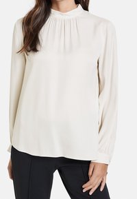 Blusa crema con maniche lunghe, colletto alto, dettagli arricciati sottili e tessuto liscio e leggero. Indossata con pantaloni neri.