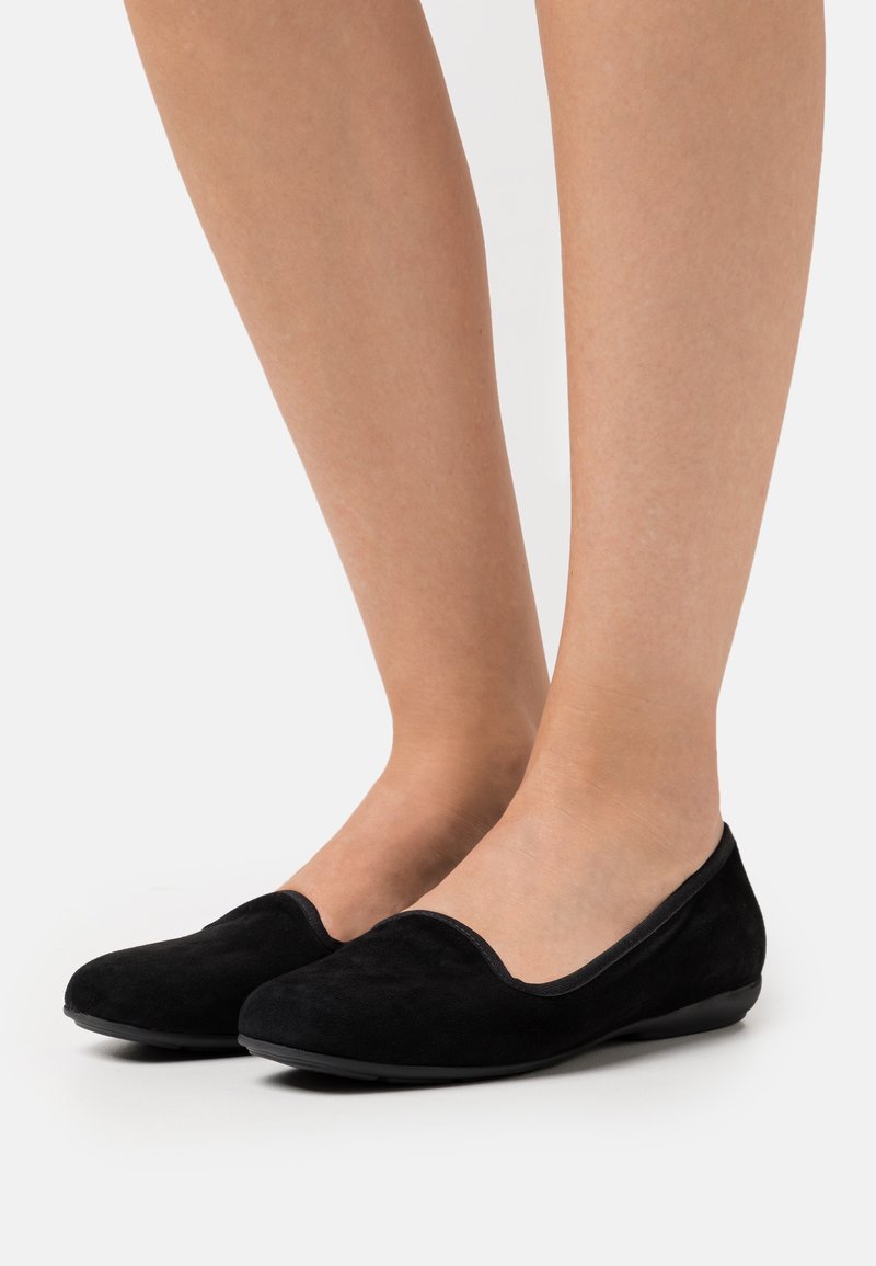 Gambe che indossano scarpe basse slip-on in suede nero, in piedi su uno sfondo bianco, che mostrano il design e la vestibilità delle scarpe.