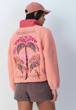 Naine kannab roosat fliisjopet tikitud palmiidega seljal, pruunide lühikeste pükstega ja tolmuroosa nokamütsiga, seisab küljepeal.