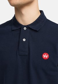 Marine polo skjorte laget av teksturert stoff, med en rød brodert logo og to hvite knapper ved kragen.