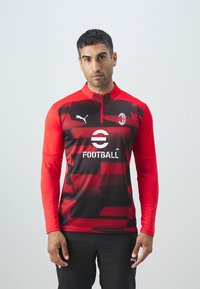 Piros és fekete, hosszú ujjú futball edzőfelső, cipzáras gallérral, terepmintával és logó kiegészítőkkel.