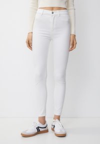 Jean skinny blanc avec une texture lisse, taille haute et fermeture à boutons. Assorti à des baskets blanches avec des accents noirs.