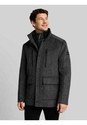 Man wearing a dark grey zip-up coat with high collar and flap pockets, standing against a plain light grey background.