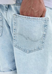 Jeans din denim albastru deschis cu un singur buzunar la spate, având un design curbat cusut. Materialul are un finisaj moale și texturat.