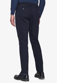 Marineblaue Hose mit Slim-Fit-Schnitt, aus glattem Material, mit Gesäßtaschen und sichtbaren Nähdetails. Kombiniert mit schwarzen Schuhen.
