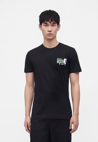Νέος άνδρας που φοράει μαύρη μπλούζα Moschino με ένα μικρό πολύχρωμο λογότυπο στο αριστερό στήθος, σε απλό λευκό φόντο.