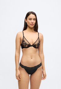 Musta läpikuultava rintaliivit, joissa kukkakirjailu, kolmionmuotoiset kupit ja ohuet olkaimet. Yhteensopiva musta pitsinen stringi, jossa aaltoilevat reunat ja rusetti.