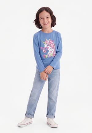 Smilende barn stående med krydsede hænder, iført en blå langærmet skjorte med en enhjørning og teksten "DREAMER", lyse jeans og hvide sneakers.