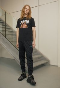 Fekete grafikás póló, amelyen a "JORDAN" felirat látható, narancssárga mez dizájnnal. Fekete sportnadrággal párosítva, kék csíkokkal, és fekete sportcipővel.