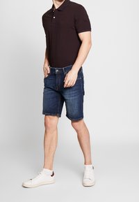 Polo de color marrón oscuro con mangas cortas, combinado con shorts de mezclilla azul oscuro hasta la rodilla y zapatillas blancas. Diseño simple, ajuste casual.
