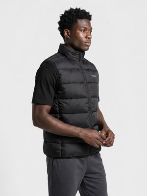Man met kort krullend haar die een zwart doorgestikt vest draagt over een zwart T-shirt en een donkere broek, staand tegen een effen lichte achtergrond.