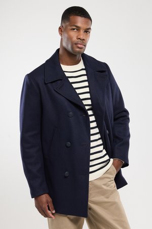 Marineblauer Wollpeacoat mit doppelter Knopfleiste, breiten Revers und zwei Knöpfen. Gestreifter Pullover darunter; das Model posiert mit einer Hand auf der Hüfte.