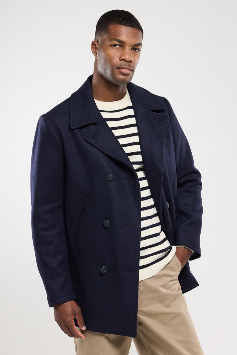 Abrigo de lana azul marino tipo peacoat con cierre de doble botonadura, amplias solapas y dos botones. Jersey a rayas debajo; el modelo posa con una mano en la cadera.