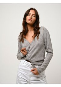 Cardigan grigio a maniche lunghe, con scollo a V e dettaglio frontale attorcigliato, indossato su jeans bianchi a vita alta. Tessuto morbido, design casual.