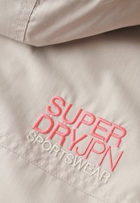 Gros plan sur un tissu beige avec du texte brodé rose et blanc « SUPER DRY JPN SPORTSWEAR » sur un vêtement de sport.