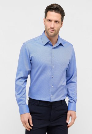 Homme portant une chemise bleue à manches longues rentrée dans un pantalon noir, debout devant un fond gris clair.