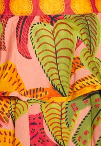 Barevná látka s tropickým vzorem listů v zelené, červené a žluté barvě na růžovém pozadí; má volánové okraje a dekorativní vyšívání.