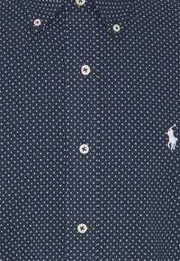 Marinblå skjorta med knappar och ett litet vitt diamantmönster, klassisk krage och kontrasterande knappar. Har en broderad logotyp på vänster bröst.