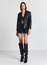 Blazer in ecopelle nera con borchie argentate, abbinato a pantaloncini neri e stivali al ginocchio. Presenta un rever con intaglio e un design corto.