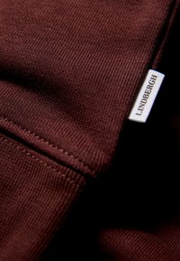 Tessuto color marrone scuro con una trama a righe; presenta un'etichetta bianca cucita sul bordo con "Lindbergh" stampato in nero.