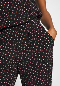 Tissu noir avec un petit motif floral rouge et violet. Le design offre une coupe décontractée avec une ceinture élastique et des poches.