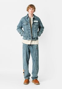 Chaqueta de mezclilla azul claro con dos bolsillos en el pecho, usada sobre una camisa beige, combinada con pantalones de mezclilla holgados que tienen un parche y cremalleras laterales. Zapatos marrones.