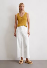 Senfgelbes Tanktop mit V-Ausschnitt und Knopfdetails, kombiniert mit hochgeschnittenen weißen weit geschnittenen Jeans und neutralen, geflochtenen Plateausandalen.