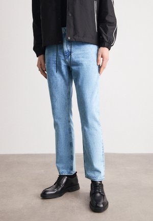 Personne portant un jean bleu clair droit, des chaussures noires à lacets et une veste noire avec un détail de bande blanche sur les manches.