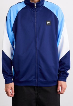 Homme portant une veste zippée bleu marine avec des blocs de couleur bleu clair et blanc sur les manches, avec un petit logo Nike Air sur la poitrine.