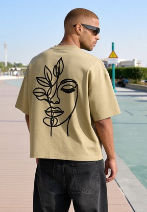 Homme portant un t-shirt beige oversize avec un design en ligne noire représentant un visage et des feuilles dans le dos, debout à l'extérieur sur un chemin pavé sous un ciel clair.