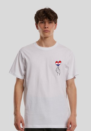Jeune homme portant un t-shirt blanc avec une main faisant un cœur avec les doigts tenant un cœur aux couleurs du drapeau croate sur la poitrine gauche.