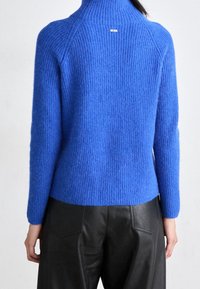 Frau in einem leuchtend blauen Rippstrickpullover mit kleinem "BOSS"-Logo im oberen Rückenbereich, kombiniert mit schwarzen Lederhosen.